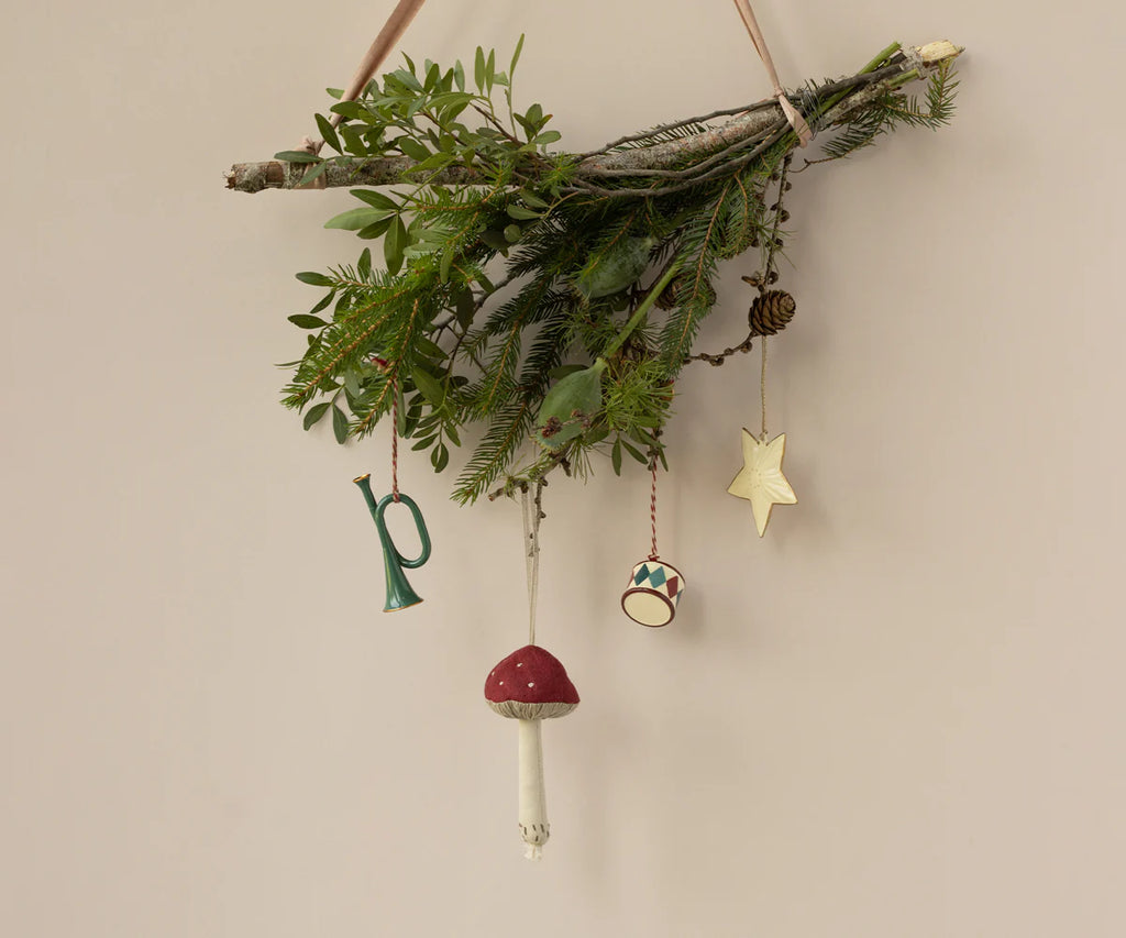 A minimalist wall hanging features an arrangement of green branches decorated with Christmas tree ornaments, including the Maileg Mushroom Ornament in red and white, a green horn, a small drum, a gold star, and a pine cone.