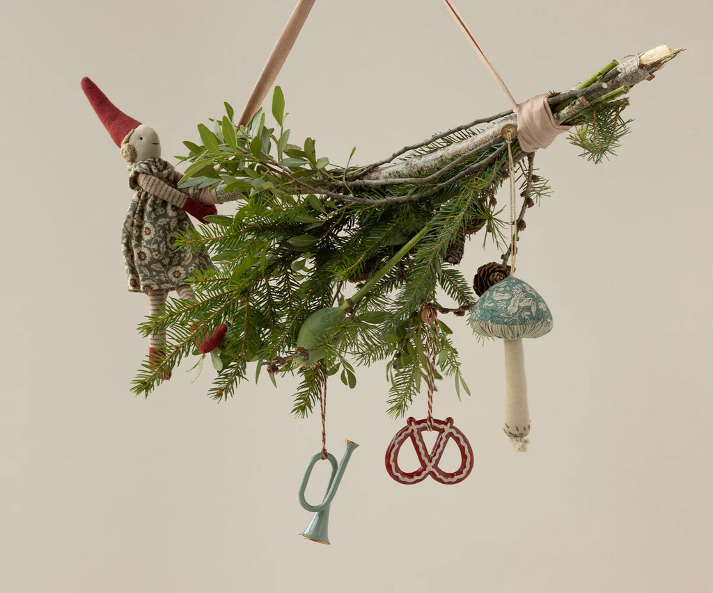 A small branch adorned with evergreen sprigs, a fabric gnome doll, and charming ornaments like the Maileg Fabric Ornament, Mushroom - Blossom - Green, plus a red pretzel, miniature blue horn, and other embroidered decorations.
