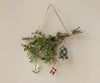 A bundle of twigs and greenery tied with a ribbon hangs on a wall, adorned with Christmas ornaments—a rocking horse, Danish flag, and the Maileg Fabric Ornament - Bell, Medium—against a neutral background.