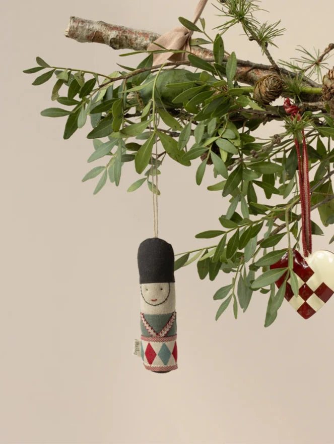 A decorative branch suspended by ribbons, adorned with green leaves and small pinecones, offers an exclusive look. From it hangs the Maileg Guard Ornament - Blue: a fabric doll with a black hat, alongside a white and red woven heart. The background is a neutral beige.