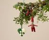 A festive scene showcases a green and white woven heart ornament accompanied by delightful Maileg ornaments. The Maileg Pixy Ornament - Boy, clad in a red outfit, hangs from a lush, leafy branch adorned with berries and pine cones, infusing whimsical charm into holiday home decor.