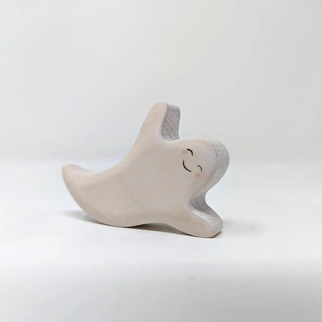 The Wooden Flying Ghost, a small, smooth, white wooden figurine hand-crafted from sustainable hardwood, stands against a plain background. This Halloween ghost has a cheerful, happy expression with its arms and one leg lifted as if dancing or floating. The minimalist design gives it a cute and friendly appearance.