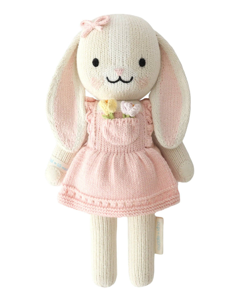 Cuddle + Kind Clover the Bunny (Blossom) is a hand-knit plush bunny in a pink flower dress with a bow on one ear, long floppy ears, black eyes, and a sweet smile—beautifully handmade in Peru under fair trade practices.