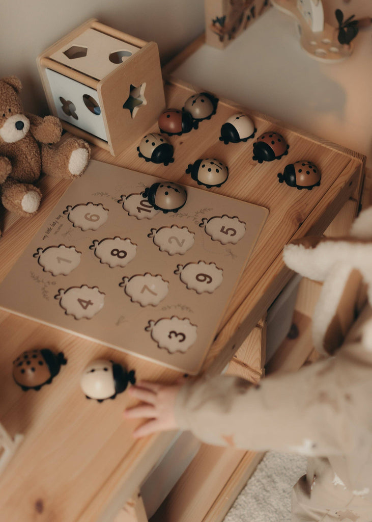 A child reaches for the My Little Lady Bug Wooden Puzzle on a small table, with a teddy bear and wooden peg puzzle shape sorter nearby.