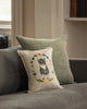 The Coral & Tusk Kitten Cameo Pillow, embroidered with a Tuxedo Cat and colorful flowers, rests against a plain green cushion on a gray sofa in a softly lit room.