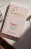 Emma | Collector's Edition | Hardcover, in pink with gold lettering and a white basket of strawberries on the cover, rests on a white surface as sunlight casts gentle shadows across the scene.