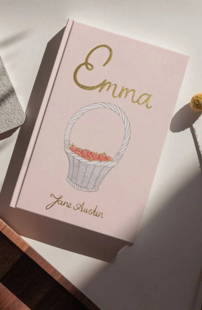 Emma | Collector's Edition | Hardcover, in pink with gold lettering and a white basket of strawberries on the cover, rests on a white surface as sunlight casts gentle shadows across the scene.