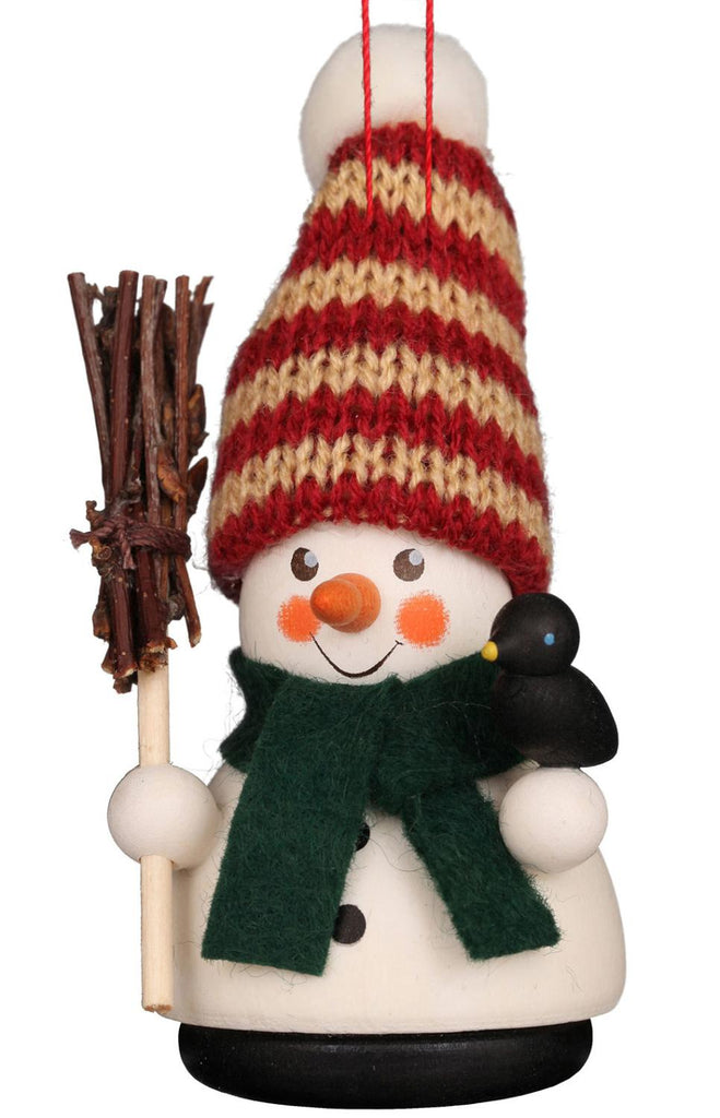 The Christian Ulbricht Ornament - Snowman showcases a hand-painted festive snowman wearing a striped red and beige knitted hat with a green scarf. It holds a bundle of twigs as a broom, with a small black bird perched on its side.