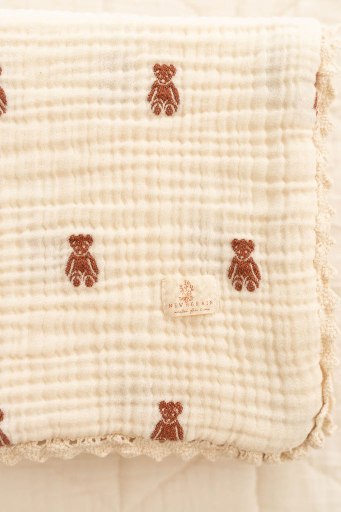 The Organic Cotton Layered Gauze Baby Blanket - Teddy (ships in one week) is a soft, cream-colored blanket with small brown teddy bear patterns, scalloped lace edging, and a sewn "NEW ORGANIC Sense of Place" label on the corner.