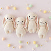 Four Cuddle + Kind Baby Bunny - Confetti dolls with floppy ears are arranged in a row on a light pink surface, surrounded by pastel eggs and small white flowers.