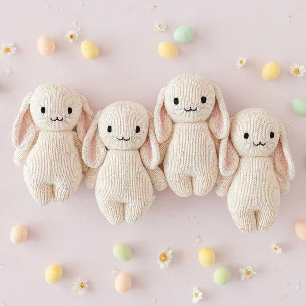 Four Cuddle + Kind Baby Bunny - Confetti dolls with floppy ears are arranged in a row on a light pink surface, surrounded by pastel eggs and small white flowers.