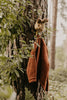 A brown sweater and rabbit mask hang from the Coat Hanger - Brown Hare, mounted on a tree trunk in a forest and partly framed by green leaves.