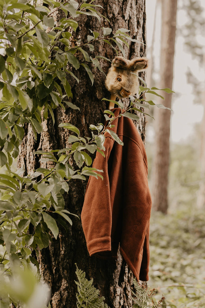 A brown sweater and rabbit mask hang from the Coat Hanger - Brown Hare, mounted on a tree trunk in a forest and partly framed by green leaves.