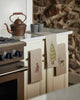 The Coral & Tusk Peek a Tree Tea Towel hangs on cream kitchen cabinets beside a stove, with a rustic teapot and two mugs on the counter and a stone wall in the background.