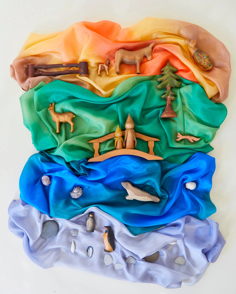 The Sarah's Silk Enchanted Playsilk in Ocean serves as the blue sea, while wooden animal figures, trees, and rocks are placed on corresponding sections of colorful draped playsilks to inspire imaginative play and demonstrate various ecosystems.