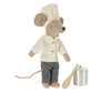 A Maileg Chef's Kitchen Starter Set toy dressed as a chef, wearing a white hat and jacket, black and white checkered pants, holding a wooden spoon, and standing next to a small metal pot.