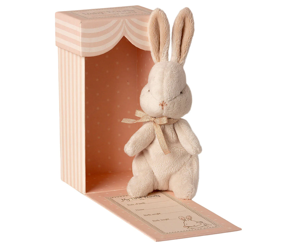 A plush beige Maileg Bunny Easter Basket Set with a bow around its neck, standing in front of a pink striped box with a pastel orange interior. The box has a bow illustration and text at the bottom
