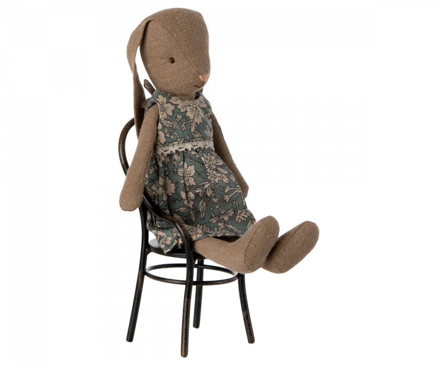 A seated Maileg Bunny, Size 1, Brown - Blossom Dress made from Maileg fabric and wearing a floral-patterned dress. The Maileg Bunny, Size 1, Brown - Blossom Dress features long ears and is positioned on an ornate black metal chair. The toy has a simple, hand-crafted appearance with a rustic, decorative style, utilizing recycled polyester for added eco-friendliness.