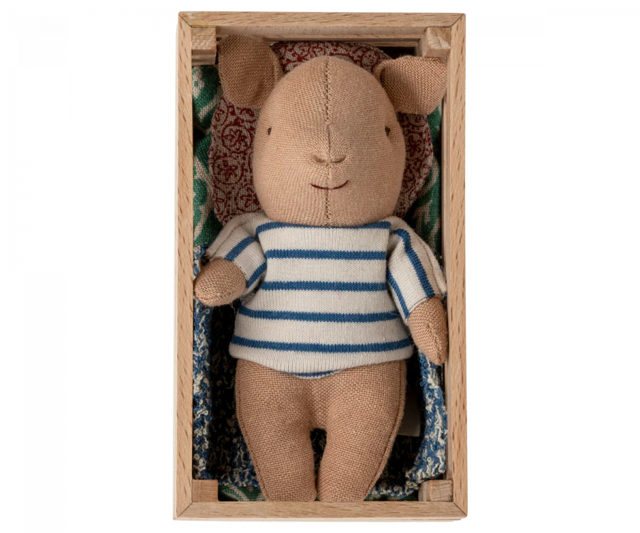 A Maileg Pig In Box, Baby - Boy with a striped blue and white shirt lies in a wooden box, resting on a patterned cushion. The bear's expression is neutral.