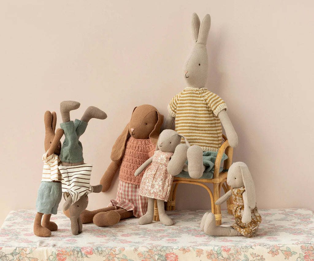 Five Maileg Bunnies, Size 3 - Knitted Shirt & Skirt, are arranged in a playful setting. One bunny is doing a handstand, another is sitting on a chair, two are standing, and one is sitting on the table. They are dressed in casual clothes made from soft natural fabrics and recycled materials. The background is a light beige wall.