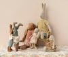 A group of Maileg Rabbit Size 3, Classic - Knitted Shirt and Pants is arranged on a floral-patterned tablecloth against a beige background. One rabbit is sitting on a wicker chair while others are standing or doing handstands. All are dressed in cute, casual clothes from a charming rabbit toy collection, made of soft natural fabrics in pastel colors.