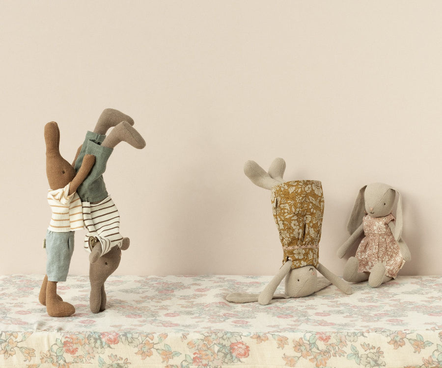 Maileg Rabbit Size 1, Chocolate Brown - Striped Blouse and Shorts toys dressed in a charming bunny wardrobe of soft natural fabrics perform playful activities on a floral-patterned surface. One Maileg rabbit, size 1, in a striped shirt helps another do a handstand, while another stands on its head next to one that sits calmly in a floral dress.