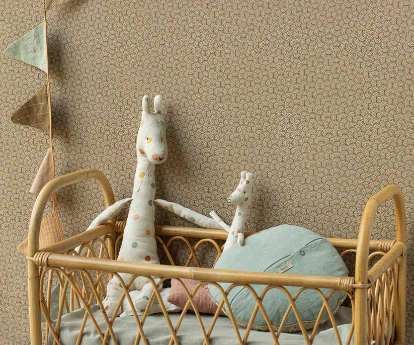 A cozy nursery corner showcasing a rattan crib with the Maileg Mini Giraffe Stuffed Animal snuggled inside and a plush giraffe toy nearby. Soft accents are provided by a round green cushion and pastel-colored bunting from the Safari Friend collection, set against a patterned wallpaper backdrop.