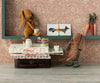 Two Maileg Bunny - Ocher, made of recycled polyester, have a tea party on a mini table stacked with books. One stands holding a teacup on top of the books, while the other sits on a small rocking chair. On the shelf above, small artworks and decorative carrots are displayed.