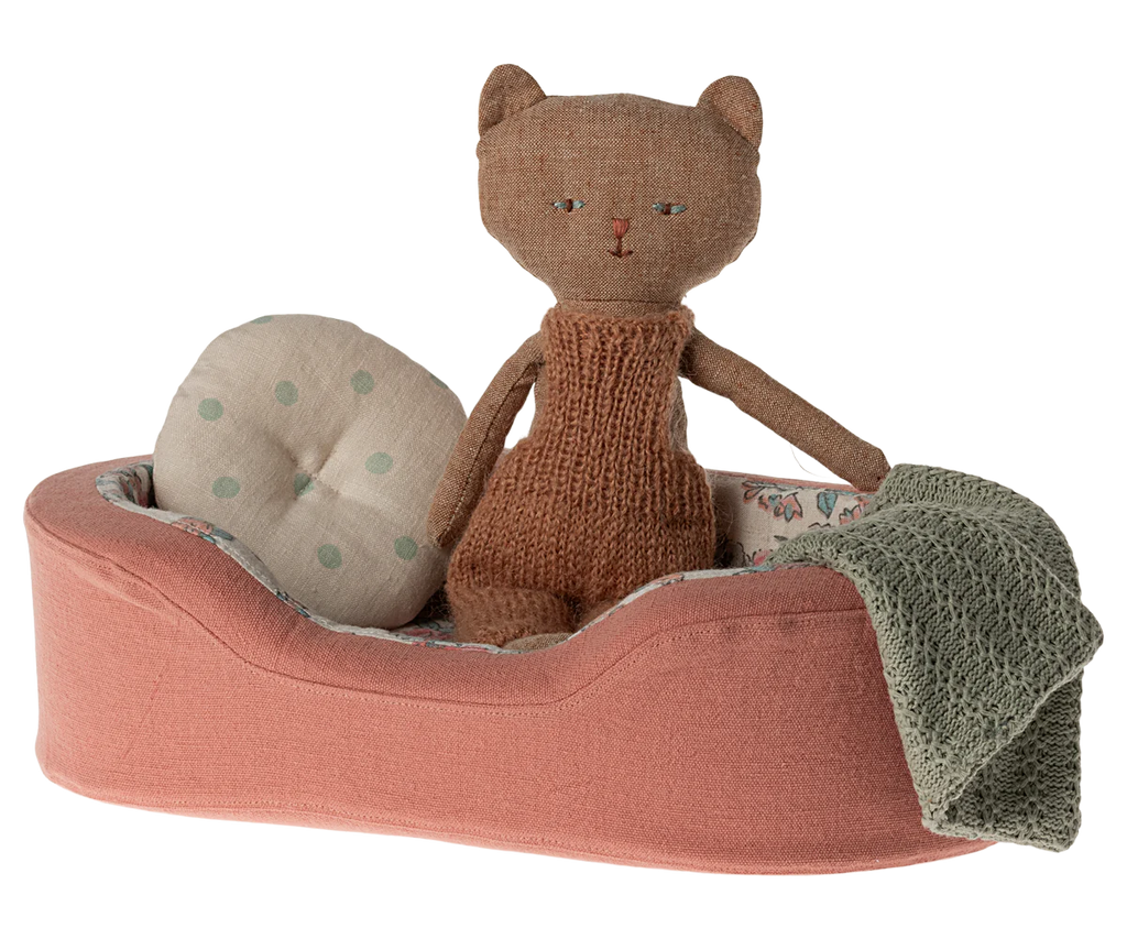 A small, stuffed toy cat wearing a knitted brown romper sits in the Maileg Small Cozy Bed - Coral. The bed features a cotton linen mattress and pillow, adorned with a beige polka dot cushion and a grey knitted blanket.