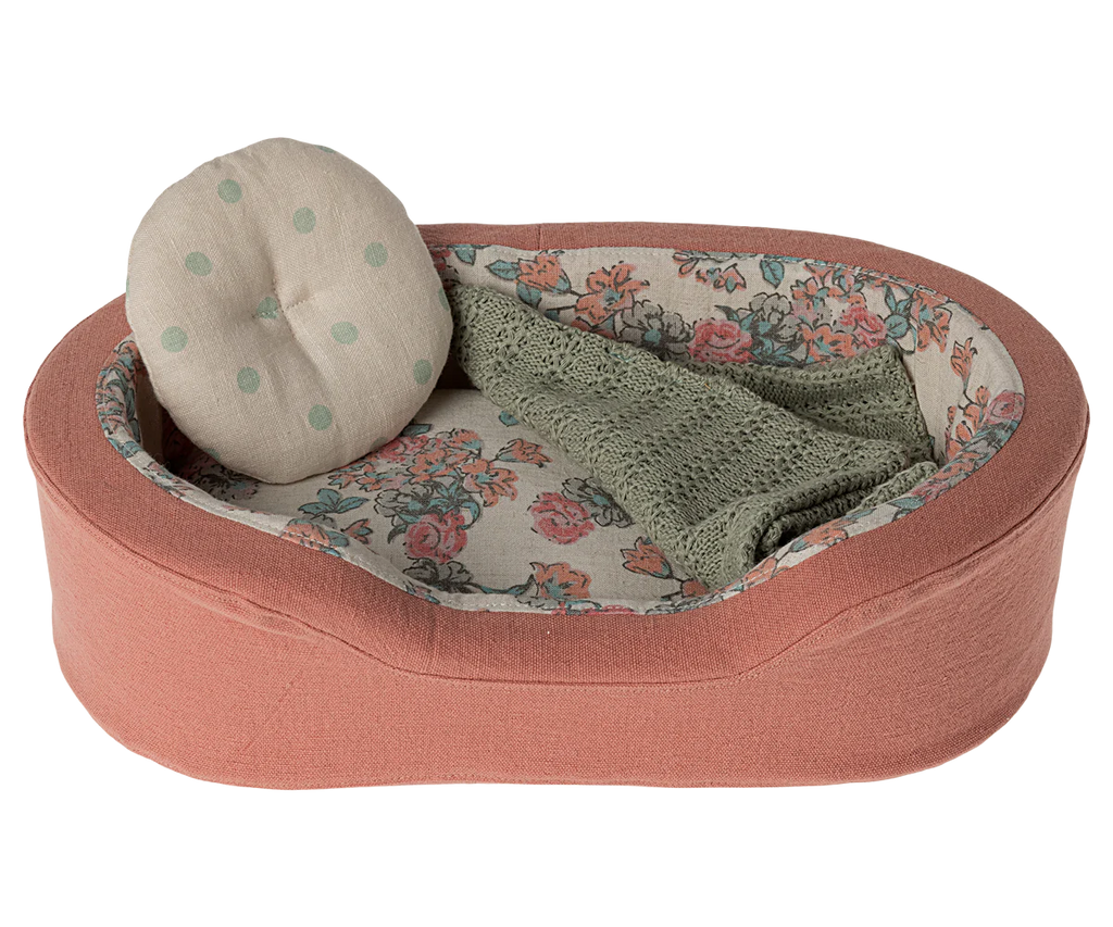 The Maileg Small Cozy Bed - Coral is a small, oval-shaped pet basket featuring a pink fabric exterior. Its interior is lined with floral-patterned fabric and includes a round cushion adorned with green polka dots, as well as a snug green knitted blanket.