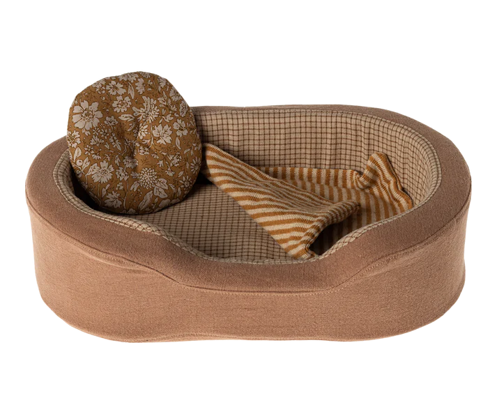 The Maileg Cozy Bed is a plush, oval-shaped pet bed featuring a beige exterior and checkered interior lining. Inside, it comes with a small, round cushion adorned with a floral pattern and a soft blanket with beige and white stripes, all neatly arranged to create a cozy space for your small pet.