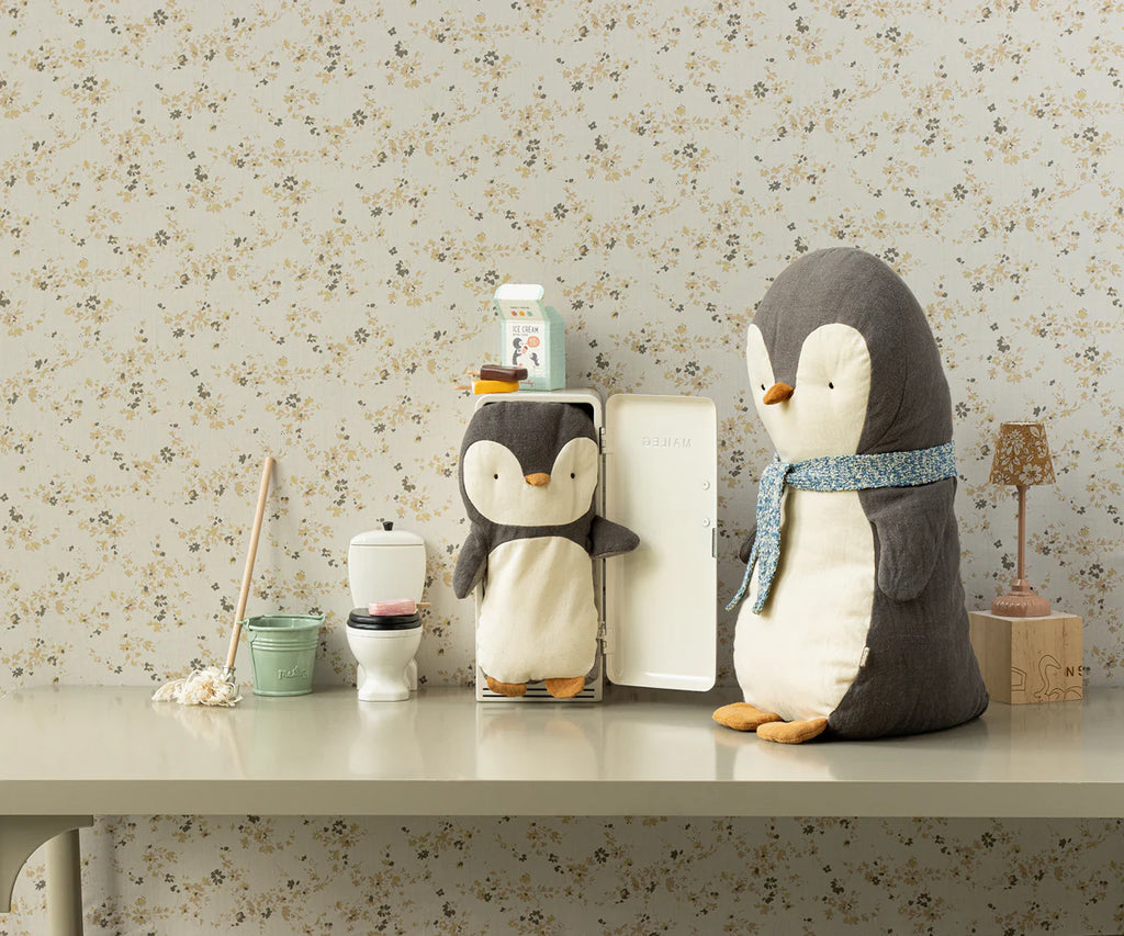 A cozy room adorned with wallpaper showcasing delicate floral patterns. On a table, plush Maileg Penguin Stuffed Animal - Small rests alongside a larger penguin amidst miniature household items: a toilet, mop, detergent, and tiny lamp. This charming setup is suitable for all ages, evoking warmth and nostalgia.