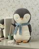 A Maileg Penguin Stuffed Animal - Medium, featuring a blue scarf, stands on a table with a miniature mop in its flipper. Encircled by cleaning accessories and a small bucket, this penguin is crafted from the softest fabrics. Another penguin peeks out from the background against a floral-patterned wall, making it ideal for all ages.