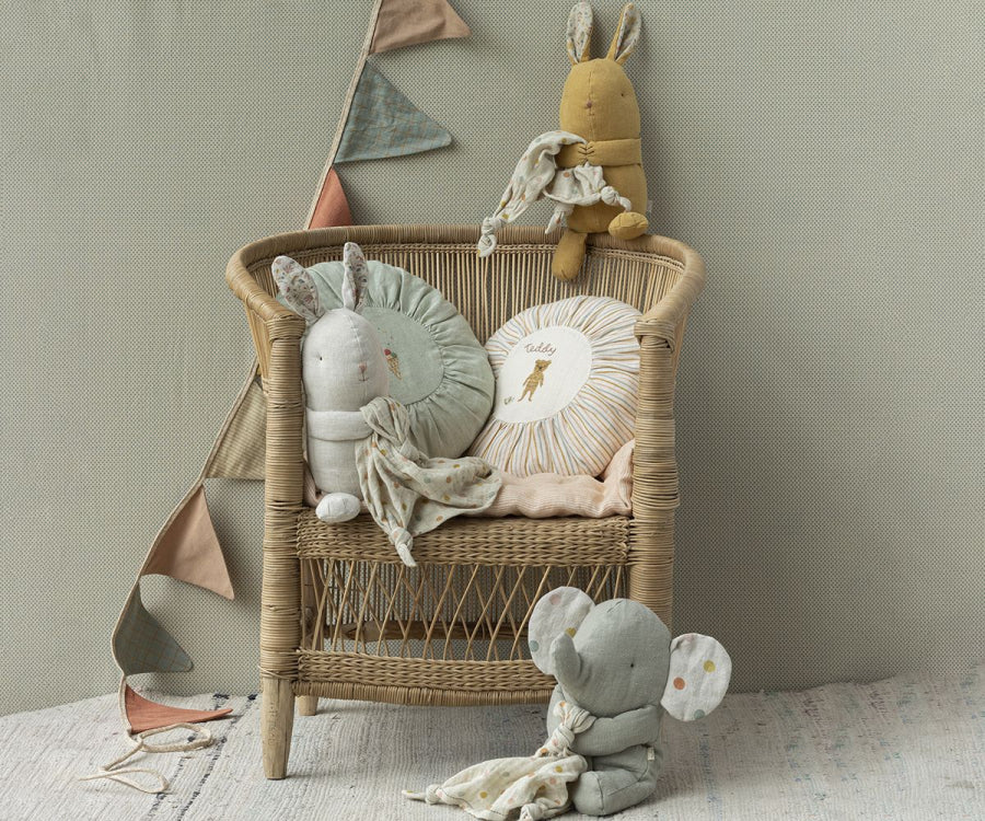 A wicker chair is adorned with soft baby toys, including a yellow rabbit, a gray bunny, and Maileg Lullaby Friends - Elephant - Iron grey (Plays Music), each holding a small blanket. A gift box sits beside the chair. Behind it, Mozart's lullaby plays softly while triangular bunting in muted colors hangs against the beige textured background.