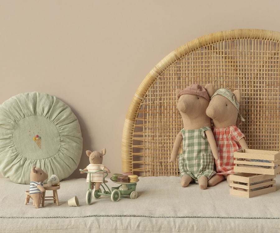 A cozy scene features two fabric mouse dolls dressed in checkered clothes, sitting on a bench with their arms around each other. Nearby, a smaller mouse plays with a wooden tricycle, and another sits at a table. A round cushion with an ice cream motif adds charm to this Maileg Pig - Boy family tableau.