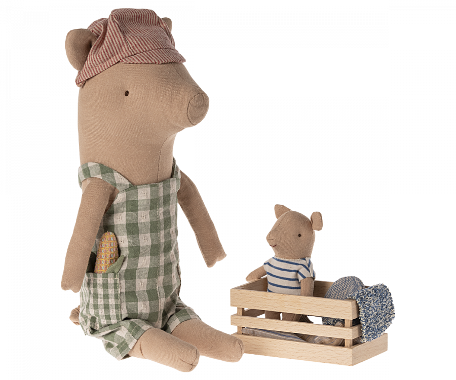 A Maileg Pig - Boy in a green checkered dress and a red striped headscarf sits with a baby bear in a striped shirt inside a small wooden crate containing two rolled-up fabric pieces, perfect for those looking to create your own pig family or add to your collection.