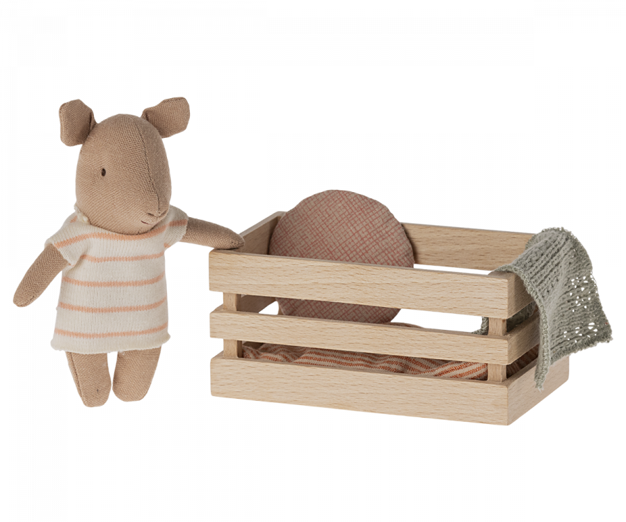 A Maileg Pig In Box, Baby Girl - Pink in a striped outfit stands beside a small wooden box containing a fabric blanket, set against a plain background. The scene conveys a cozy, playful setting.