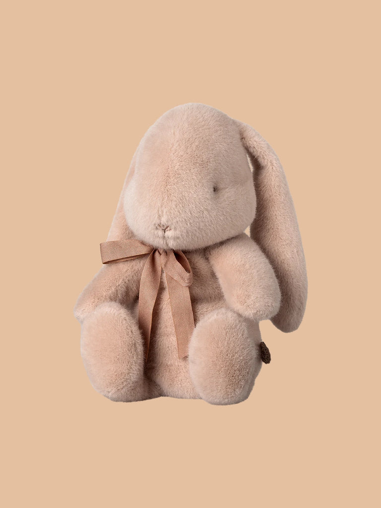 The Maileg Plush Bunny in Light Powder, available for pre-order and shipping in February, features long ears and a brown ribbon necktie. Crafted from soft plush fabric with polyester fillings, this irresistibly fluffy and cuddly bunny sits against a plain beige background.