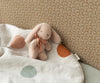 The Maileg Mini Plush Bunny - Powder features soft, floppy ears and a ribbon around its neck, nestled against a patterned background. Made from plush fabric and polyester fillings, it is snugly tucked under a blanket adorned with large pastel green and orange spots.