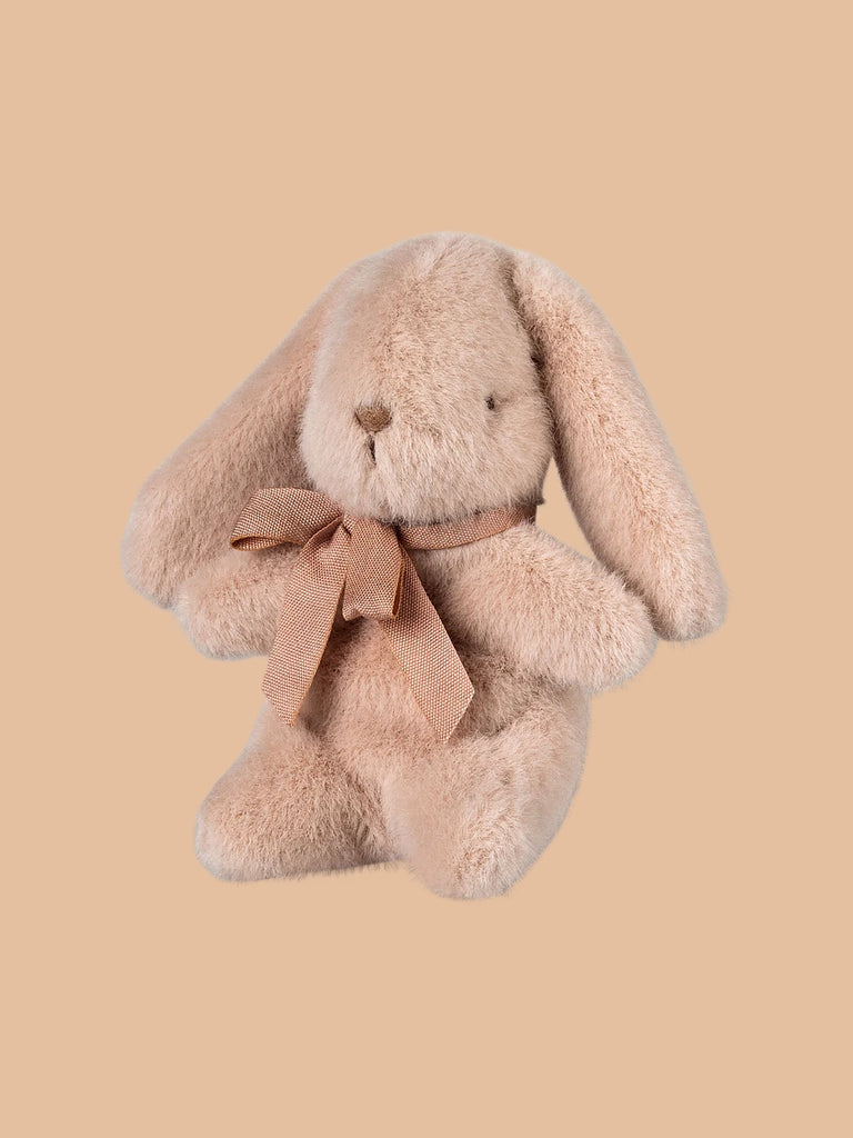 A cuddly, plush bunny in light powder color made from recycled materials has long ears and a neck ribbon, all set against a beige background. Available for pre-order as Maileg Plush Bunny, shipping late February.