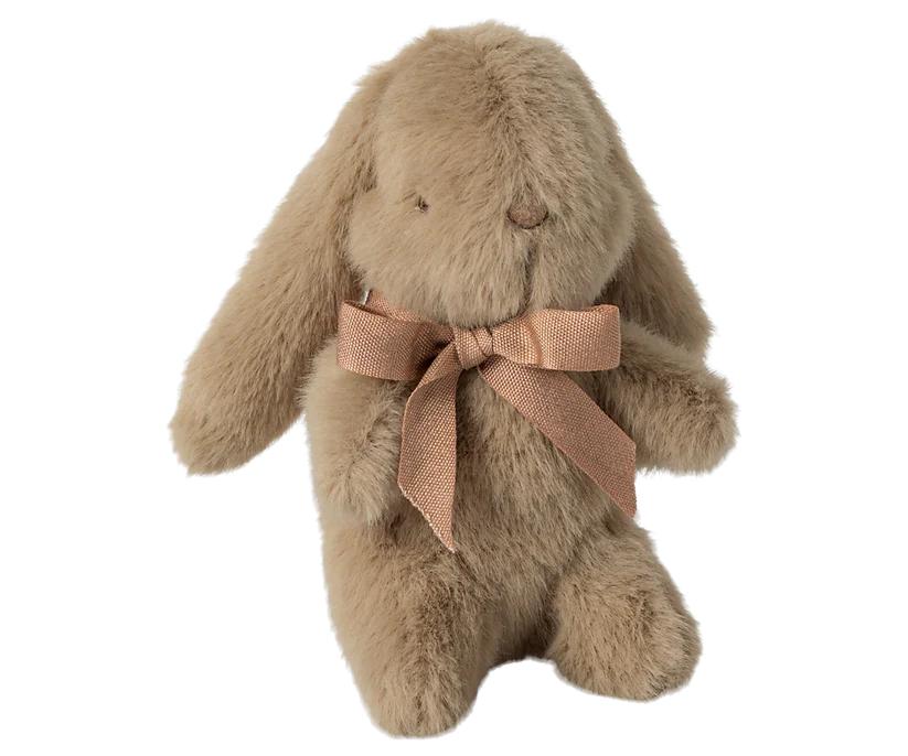 The Maileg Mini Plush Bunny in Dusty Brown is sitting upright, featuring long, floppy ears reminiscent of a plush bunny. Crafted from unique plush fabric, it is filled with polyester fillings for a soft and fluffy texture and adorned with a light brown ribbon tied in a bow around its neck.