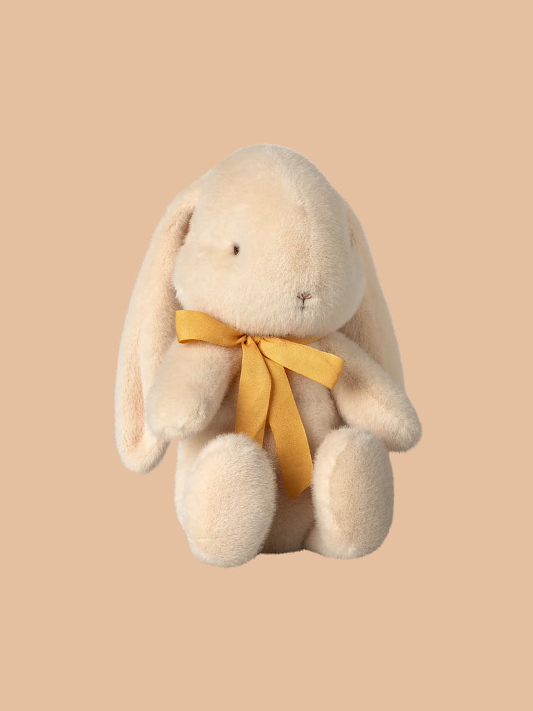 The PRE-ORDER: Maileg Plush Bunny - Medium Cream sits upright against a beige background, crafted from recycled polyester. This soft toy features a light cream hue with a yellow bow around its neck, highlighting its adorable charm.