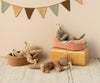 Four Maileg Plush Kittens, Cream peach (ships in approximately one week), two grey and two brown, each holding a small toy giraffe, are posed on various objects including a pink cushion, a brown basket, and a yellow box. In the background, a string of colorful triangular flags is strung across the wall.