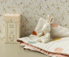 A soft, plush Maileg My First Bunny in Dusty Rose is seated on a dotted baby blanket, accompanied by a decorative box labeled "My First Bunny." This charming scene makes an ideal baby shower gift, set against the backdrop of gentle, floral-patterned wallpaper.