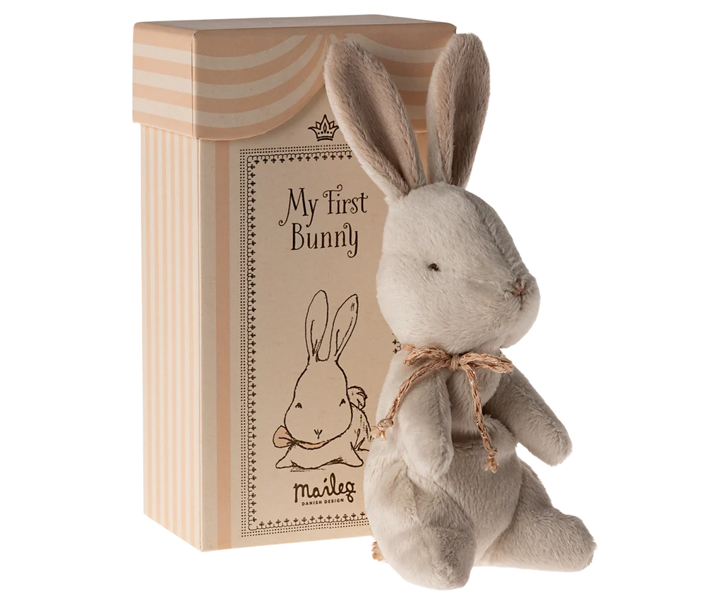 A small, soft grey plush bunny with a brown bow sits in front of a beige box with pink stripes. The gift box features an illustration of a bunny and the words "My First Bunny" along with the brand name "Maileg My First Bunny - Dusty Rose," making it an ideal baby shower gift.