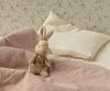 The Maileg My First Bunny - Brown, a soft plush toy with a ribbon, sits on a bed with pink checkered blanket and cream pillows against floral wallpaper—arriving in a beautiful gift box perfect for any special occasion.