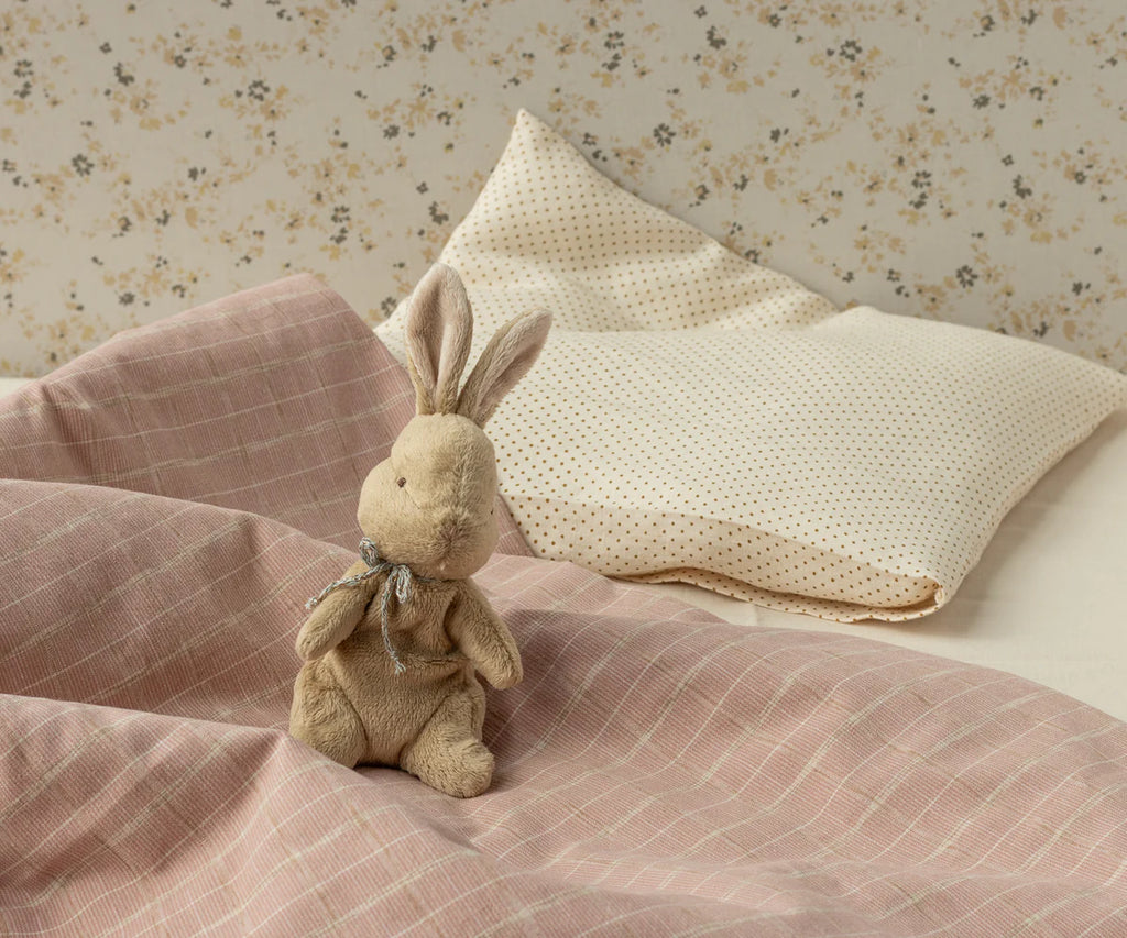 The Maileg My First Bunny - Brown, a soft plush toy with a ribbon, sits on a bed with pink checkered blanket and cream pillows against floral wallpaper—arriving in a beautiful gift box perfect for any special occasion.