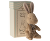 A cozy brown plush bunny with a silver ribbon sits before a “My First Bunny” gift box by Maileg, which displays an illustration of a bow-wearing bunny.