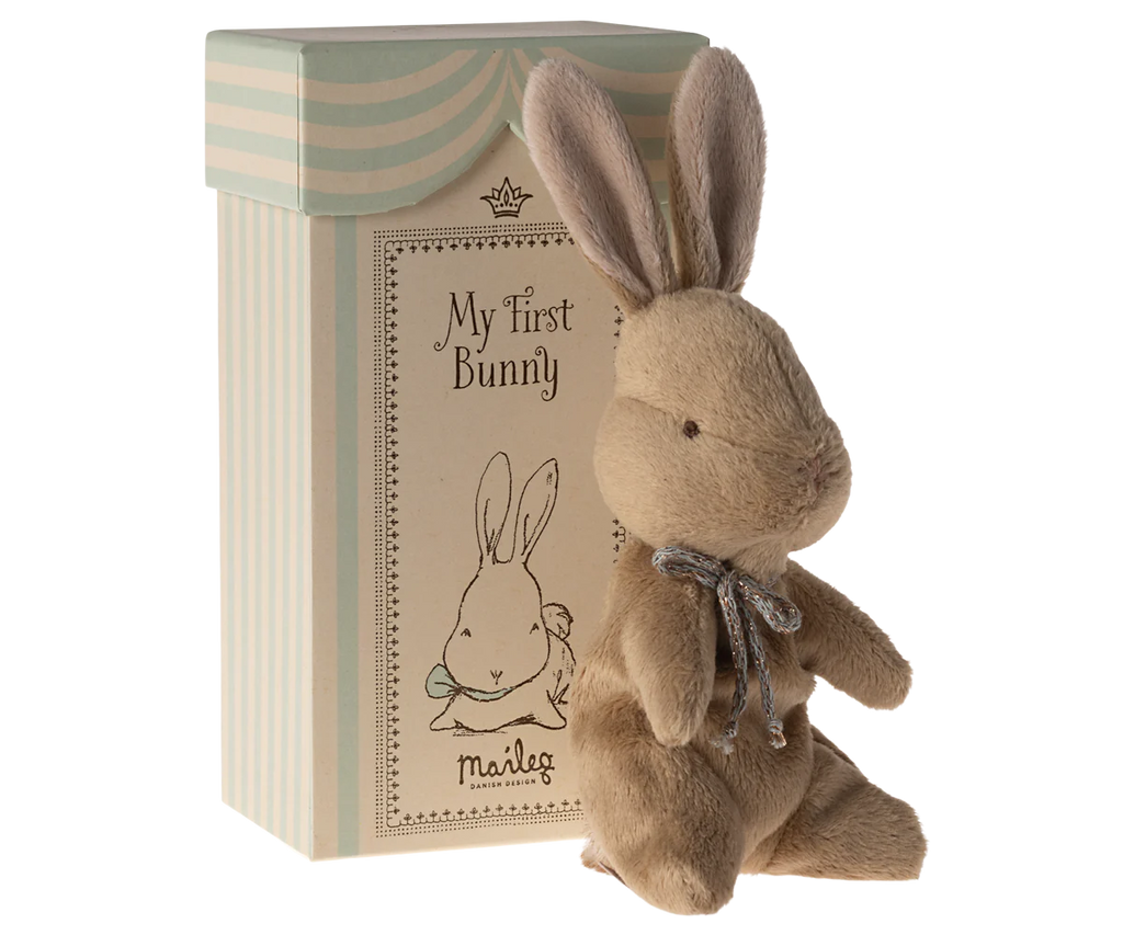 A cozy brown plush bunny with a silver ribbon sits before a “My First Bunny” gift box by Maileg, which displays an illustration of a bow-wearing bunny.