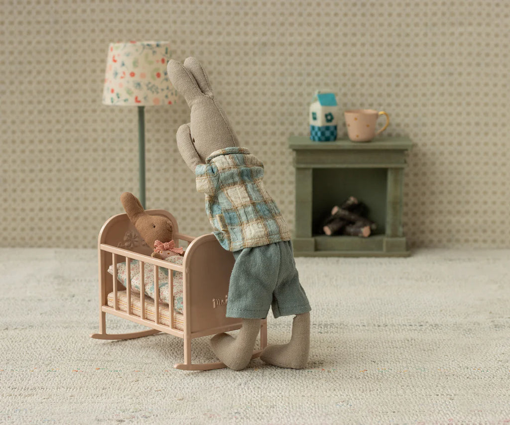 A Maileg Rabbit, Size 1 - Classic - Shirt and Shorts kneels beside a crib, comforting a smaller stuffed rabbit in a cozy, vintage-style room with miniature furniture, a lamp, fireplace, mug, and playful decor.